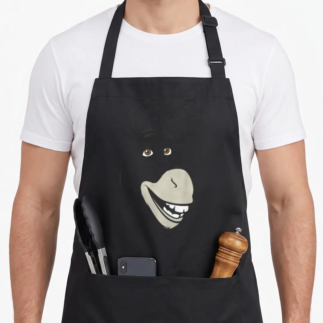 Donkey face apron - Cartoon donkey head facing right in grey, brown, and white with a wide toothy grin — shown on a black cotton canvas apron against a white background