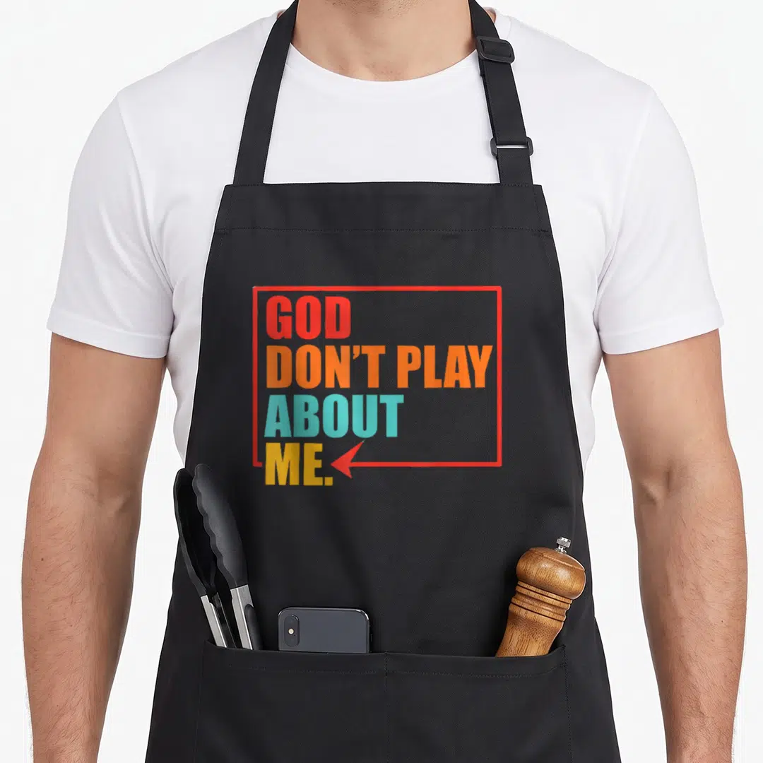 God play apron - Stacked text GOD DON'T PLAY ABOUT ME in red orange teal yellow with arrow — shown on a black cotton canvas apron against a white background