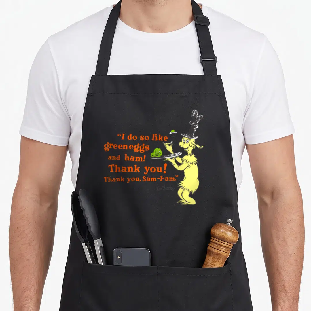 Ham green egg apron - Sam-I-Am holding green food plate next to orange quote text — shown on a black cotton canvas apron against a white background