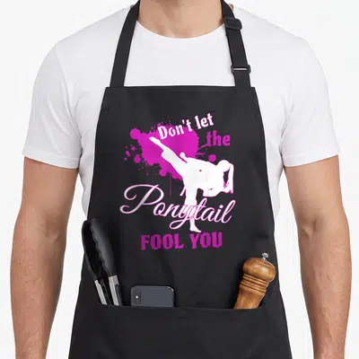 Ponytail fool apron - White martial artist silhouette kicking over magenta splatter with text Don't let the Ponytail — shown on a black cotton canvas apron against a white background