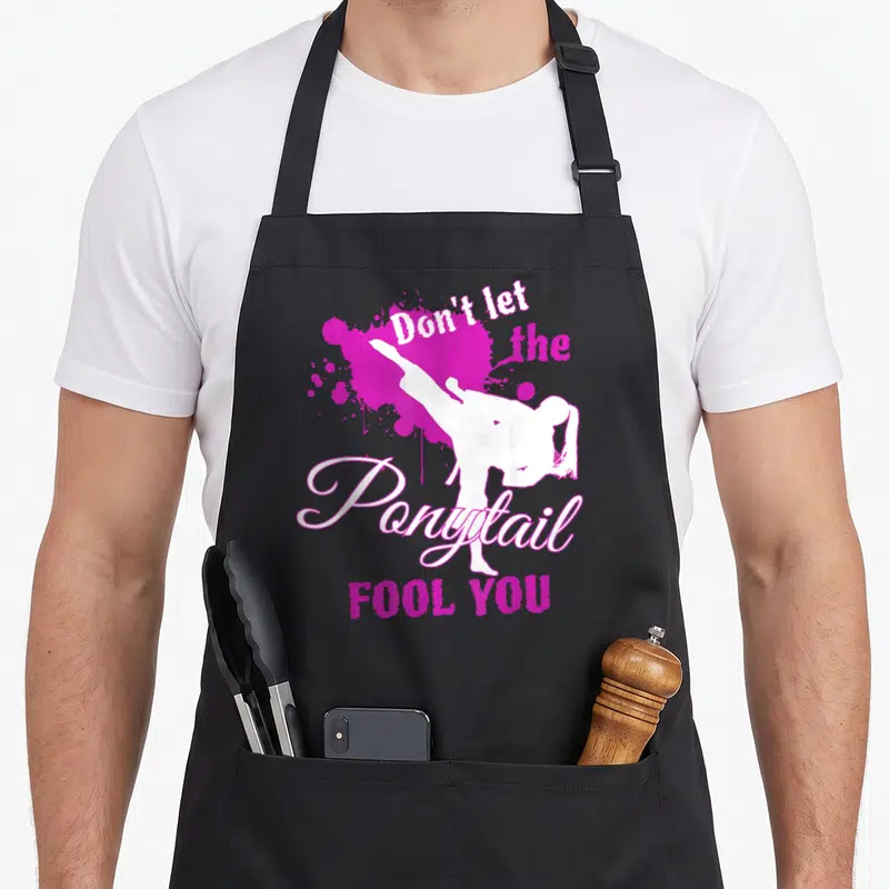 Ponytail fool apron - White martial artist silhouette kicking over magenta splatter with text Don't let the Ponytail — shown on a black cotton canvas apron against a white background