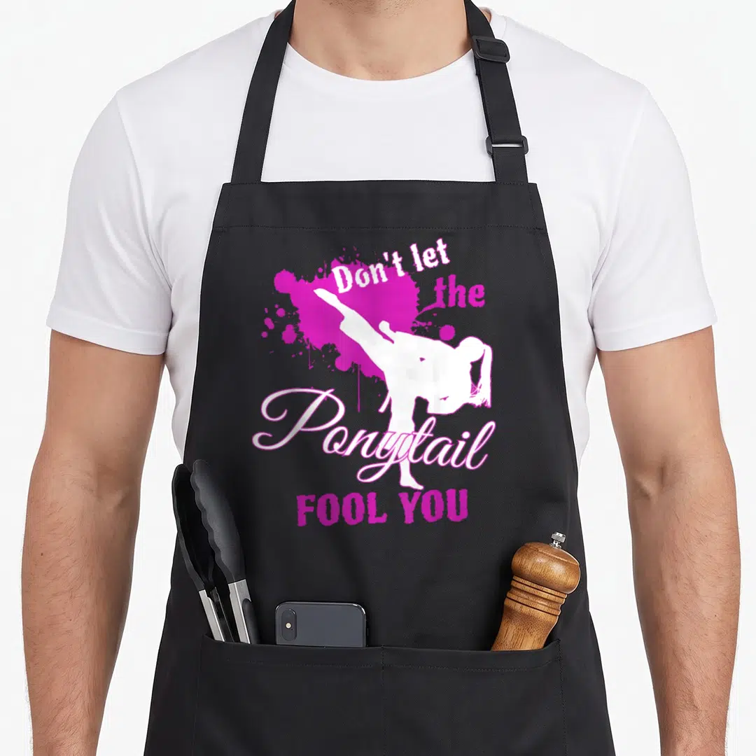 Ponytail fool apron - White martial artist silhouette kicking over magenta splatter with text Don't let the Ponytail — shown on a black cotton canvas apron against a white background