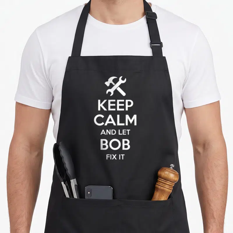 Bob fix birthday apron - White crossed hammer wrench and stacked text reading Keep Calm And Let Bob Fix It — shown on a black cotton canvas apron against a white background