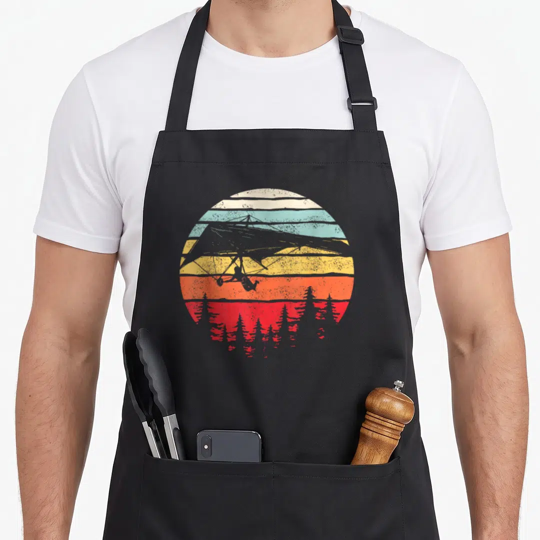 Hang crazy canary apron - Retro circular sunset with hang glider silhouette and striped colors — shown on a black cotton canvas apron against a white background