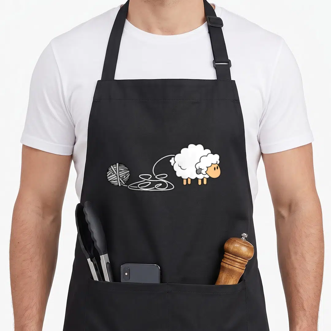 Sheep knitting apron - Cartoon sheep with white wool and peach face pulling yarn to a ball — shown on a black cotton canvas apron against a white background