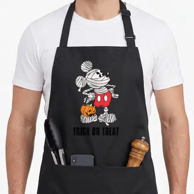 Vintage halloween mummy and me aprons - Mickey Mouse mummy holding orange pumpkin bucket with trick or treat text below — shown on a black cotton canvas apron against a white background