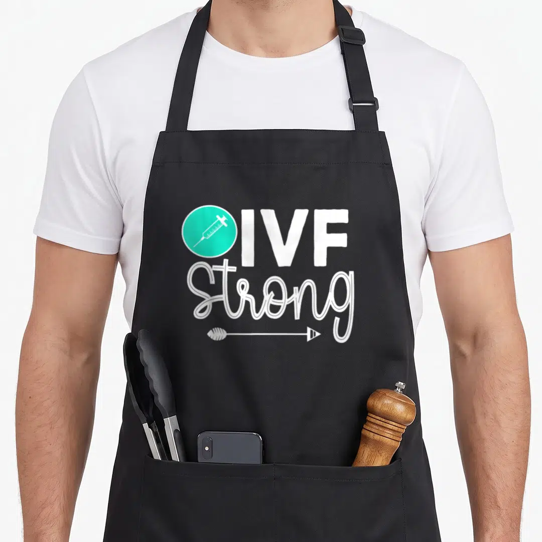 Vitro infertility awareness apron - Teal circle with white syringe next to IVF Strong text and arrow underline — shown on a black cotton canvas apron against a white background