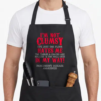 Clumsy parkinsons awareness apron - Red and white text reading I'm not clumsy it's just the floor hates me Parkinson's — shown on a black cotton canvas apron against a white background