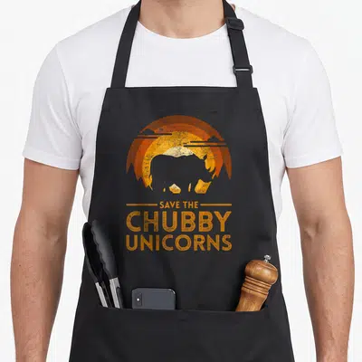 Funny save unicorn apron - Retro sunset graphic with silhouette and text SAVE THE CHUBBY UNICORNS in orange and brown — shown on a black cotton canvas apron against a white background