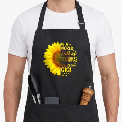 Gigi grandma's aprons - Half sunflower and text forming circle with small hearts in yellow and brown — shown on a black cotton canvas apron against a white background