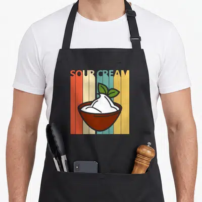 Cute sour cream aprons - Retro brown bowl with white cream and green leaves over five colorful vertical stripes — shown on a black cotton canvas apron against a white background