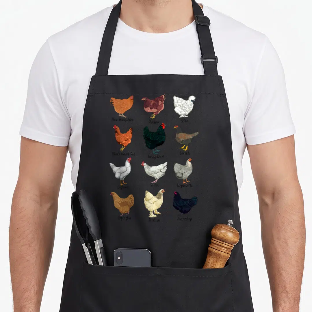 Various chicken apron - Twelve chicken breeds in a grid with labels including New Hampshire, Buckeye, and Silkie — shown on a black cotton canvas apron against a white background