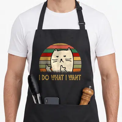 Cat apron gift - Grumpy white cat raising paws against retro stripes with orange text I DO WHAT I WANT — shown on a black cotton canvas apron against a white background