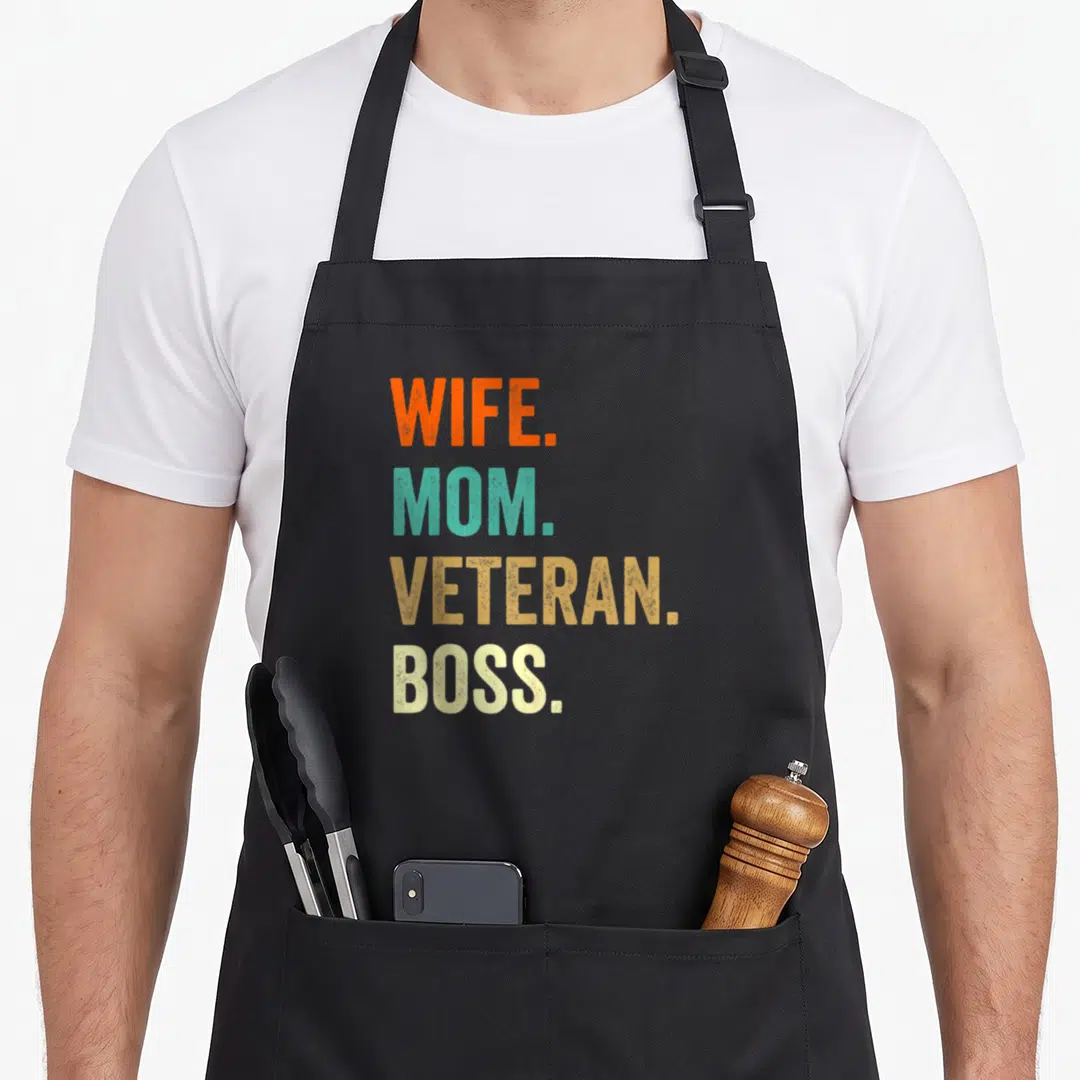 Military veteran wife apron - Stacked WIFE. MOM. VETERAN. BOSS. text in orange teal gold and cream colors — shown on a black cotton canvas apron against a white background