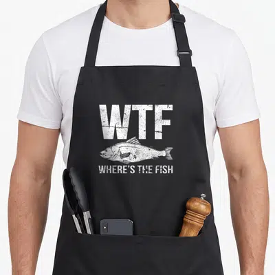 Cute fish apron - WTF Where's The Fish white distressed text above a fish sketch — shown on a black cotton canvas apron against a white background