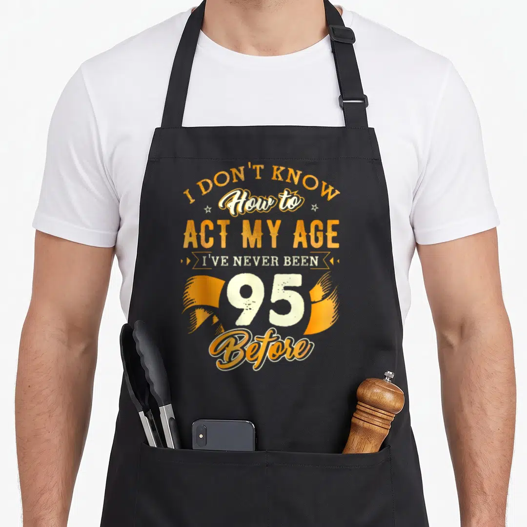 95th act birthday apron - Orange and white typography reading I Don't Know How to Act My Age 95 Before with ribbon — shown on a black cotton canvas apron against a white background
