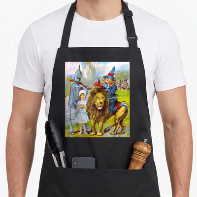 Emerald dorothy apron - Cartoon lion with scarecrow rider, tin man, girl, and castle in blue green gold white — shown on a black cotton canvas apron against a white background