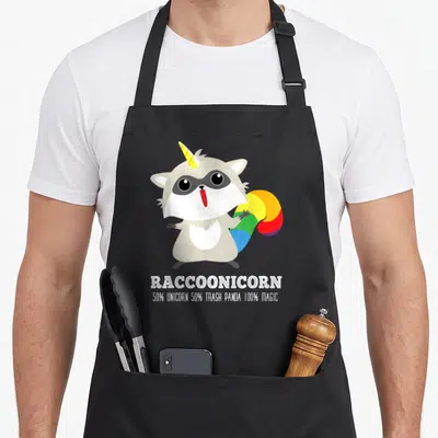 Racoonicorn unicorn apron - Grey cartoon raccoon with yellow horn and rainbow tail above white text reading racoonicorn — shown on a black cotton canvas apron against a white background