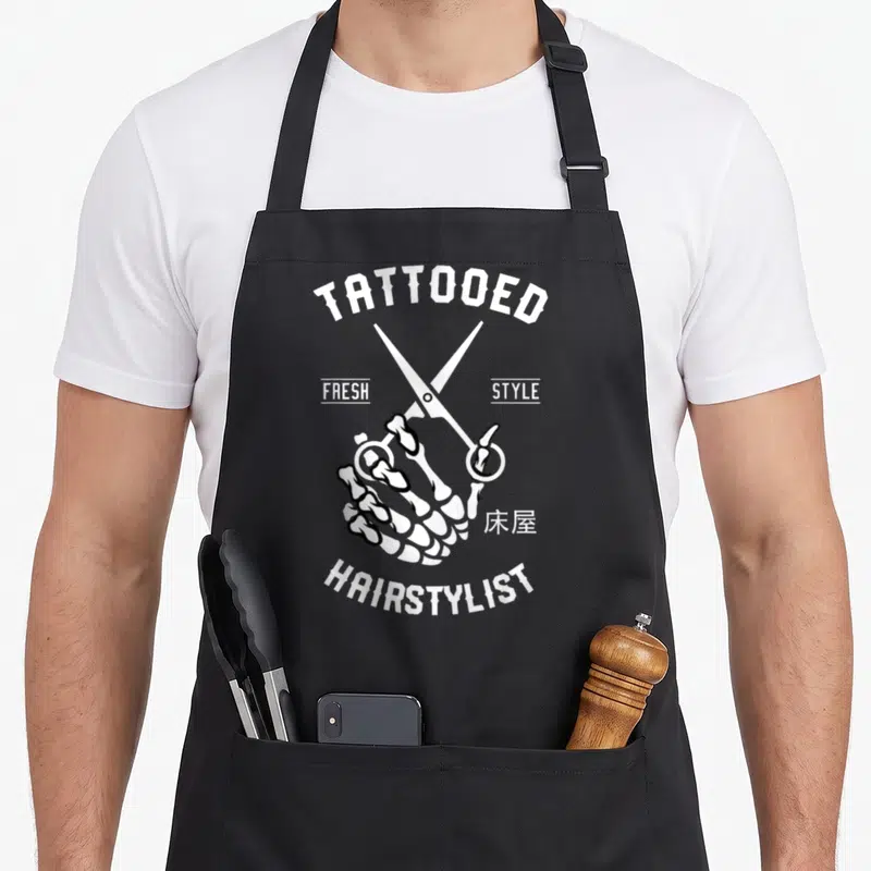 Tattooed hairstylist apron - White skeleton hand holding scissors with tattoo-style text TATTOOED FRESH STYLE — shown on a black cotton canvas apron against a white background