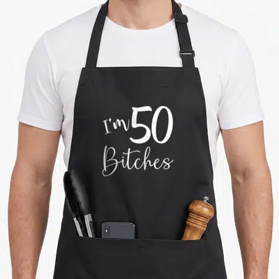 50th age birthday apron - White I'm 50 Bitches script typography on black background — shown on a black cotton canvas apron against a white background