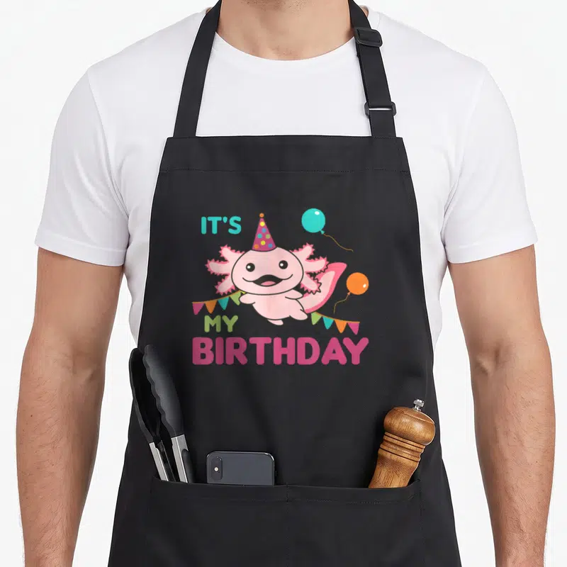 Axolotl balloon apron - Pink axolotl wearing party hat with It's My Birthday text and colorful balloons — shown on a black cotton canvas apron against a white background