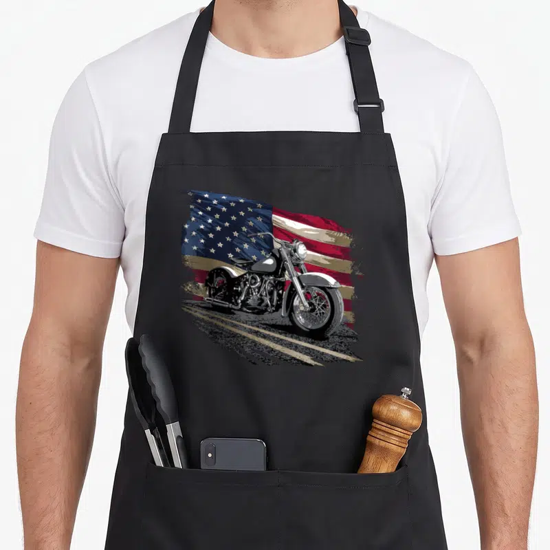Open american flag apron - Realistic motorcycle on road with waving red white blue flag background — shown on a black cotton canvas apron against a white background