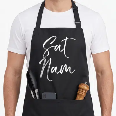 Sat grill instructor apron - White Sat Nam typography stacked vertically in a handwritten script style — shown on a black cotton canvas apron against a white background