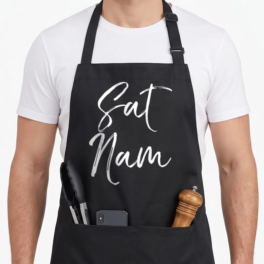 Sat grill instructor apron - White Sat Nam typography stacked vertically in a handwritten script style — shown on a black cotton canvas apron against a white background