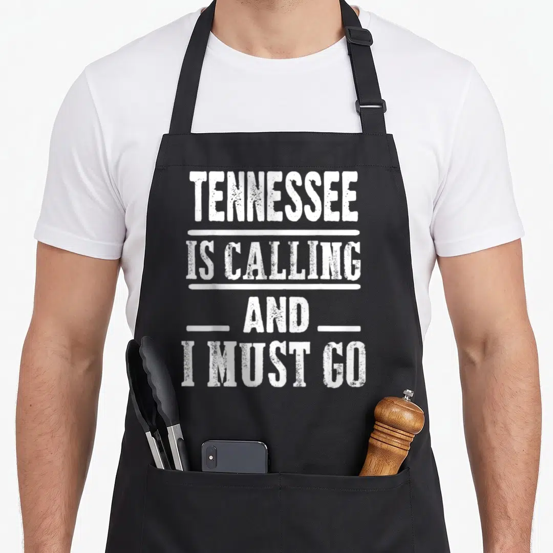 Traveling housewarming apron - White stacked text reading Tennessee Is Calling And I Must Go on black fabric — shown on a black cotton canvas apron against a white background