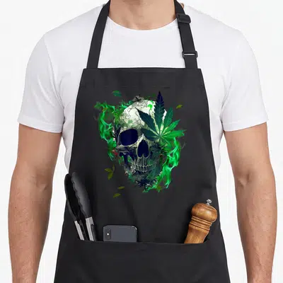 Marijuana sugar skull apron - Realistic skull covered in green cannabis leaves and swirling smoke on black fabric — shown on a black cotton canvas apron against a white background