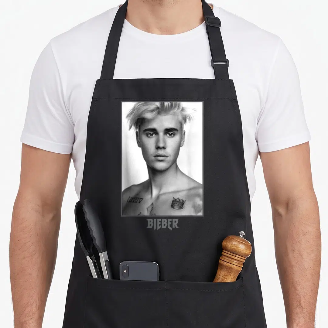 Justin sorry apron - Black and white portrait of Justin Bieber with tattoos inside a white frame and BIEBER text below — shown on a black cotton canvas apron against a white background