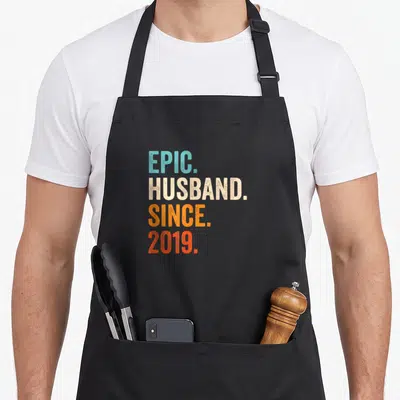 7th wedding apron - Stacked EPIC HUSBAND SINCE 2019 text in blue beige orange and red distressed font — shown on a black cotton canvas apron against a white background