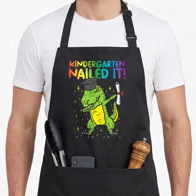 Kindergarten nailed dinosaur apron - Rainbow text reading KINDERGARTEN NAILED IT above a green dinosaur wearing — shown on a black cotton canvas apron against a white background