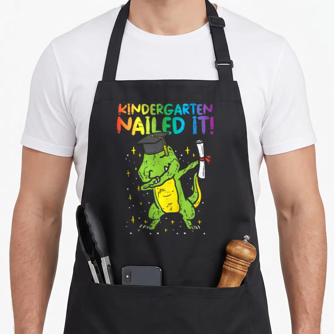 Kindergarten nailed dinosaur apron - Rainbow text reading KINDERGARTEN NAILED IT above a green dinosaur wearing — shown on a black cotton canvas apron against a white background
