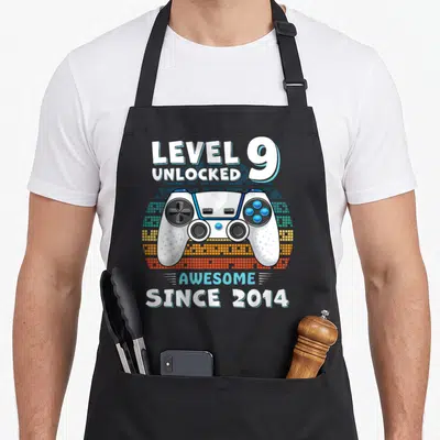 Nine birthday apron - Game controller with LEVEL 9 UNLOCKED text over pixelated background in white blue orange — shown on a black cotton canvas apron against a white background