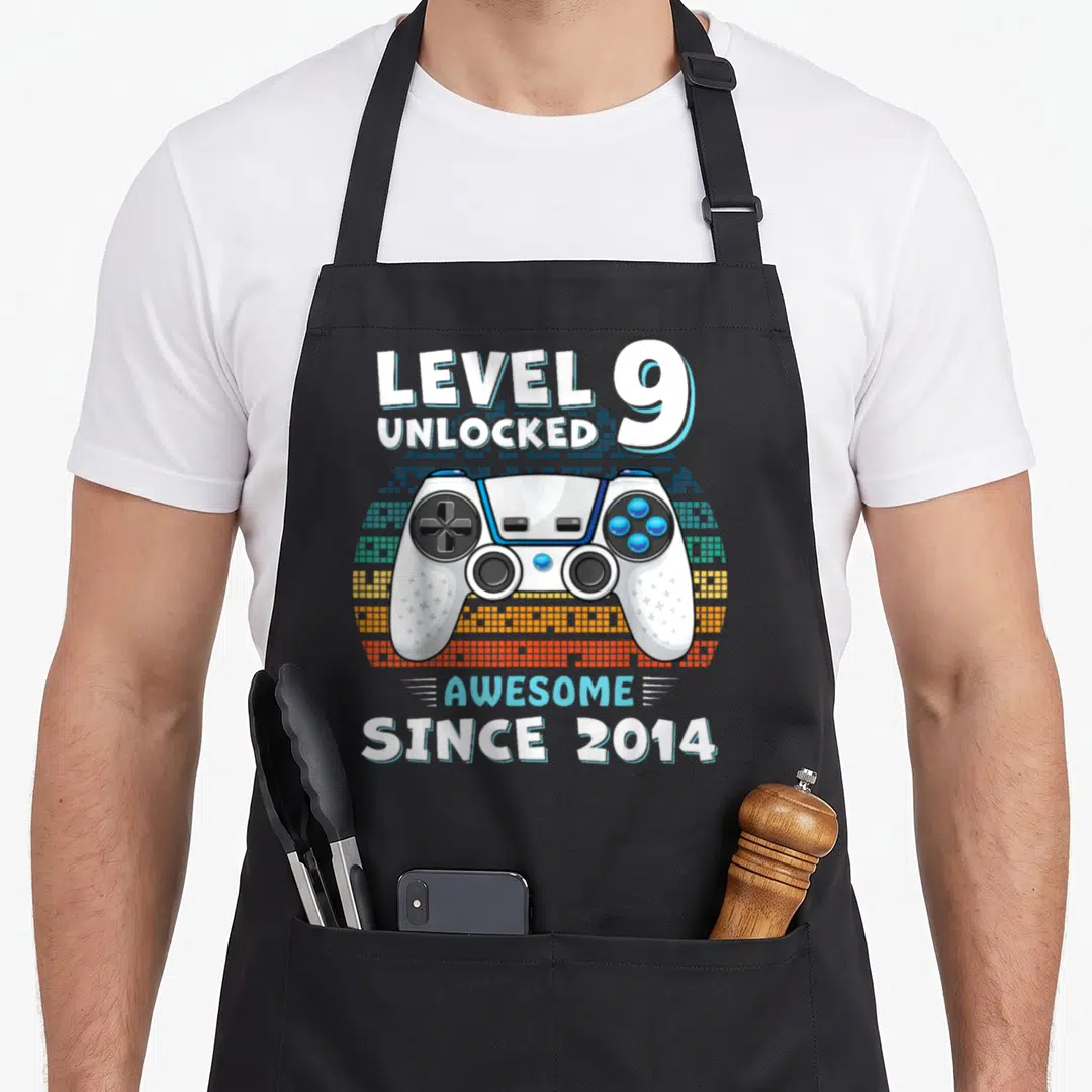 Nine birthday apron - Game controller with LEVEL 9 UNLOCKED text over pixelated background in white blue orange — shown on a black cotton canvas apron against a white background