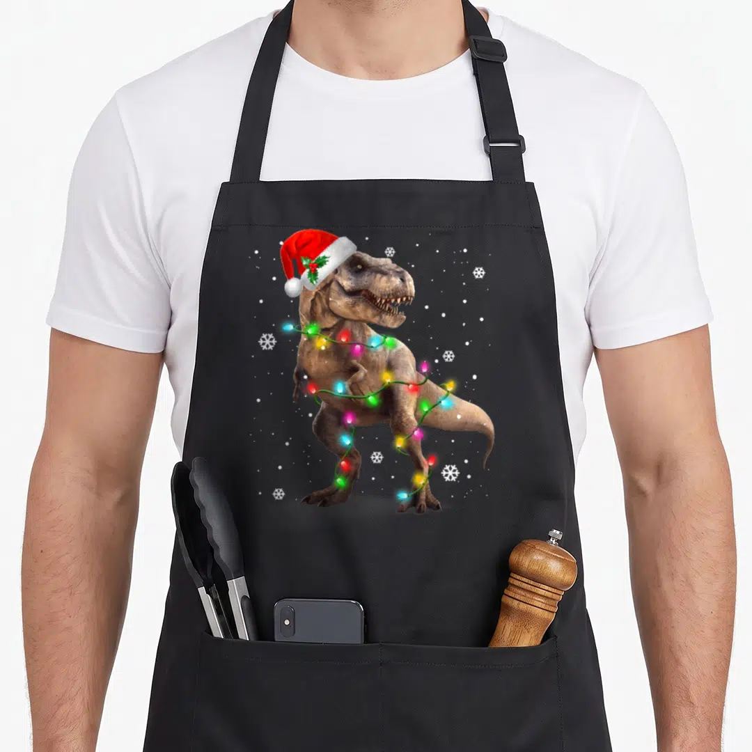Dinosaur pet apron - Realistic T-Rex wearing red Santa hat with holly, wrapped in multi-colored Christmas lights — shown on a black cotton canvas apron against a white background