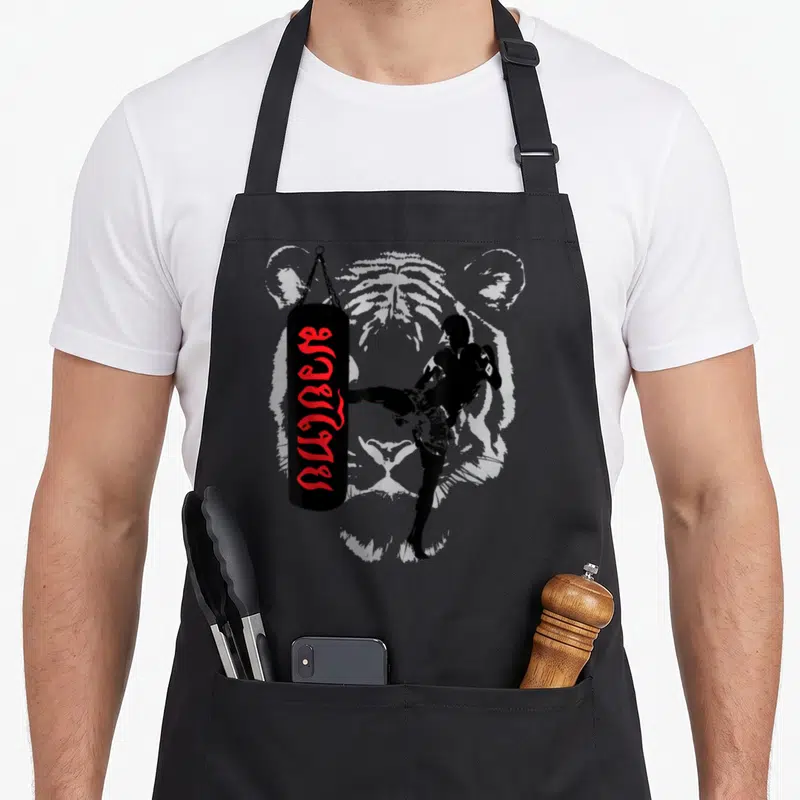 Muay thai boy apron - Fighter silhouette kicking black bag with red Thai script and grey tiger background — shown on a black cotton canvas apron against a white background