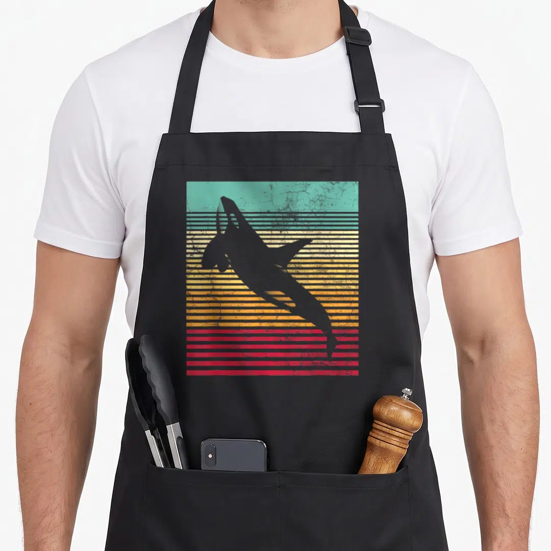 Funny whale orca dad apron - Grey shark silhouette over retro teal yellow orange and red horizontal stripes — shown on a black cotton canvas apron against a white background
