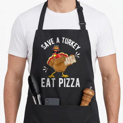 Save vegan apron - Cartoon turkey with sunglasses holding pizza box with text SAVE A TURKEY EAT PIZZA — shown on a black cotton canvas apron against a white background