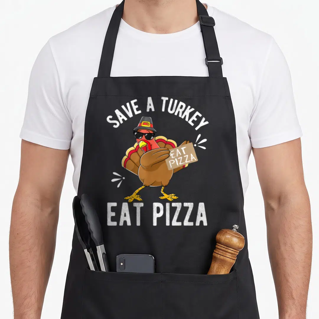 Save vegan apron - Cartoon turkey with sunglasses holding pizza box with text SAVE A TURKEY EAT PIZZA — shown on a black cotton canvas apron against a white background
