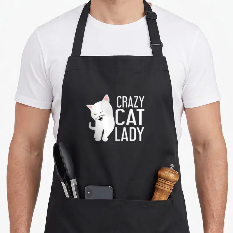 Funny costume crazy cat lady apron - White cat illustration beside stacked text reading CRAZY CAT LADY in white font — shown on a black cotton canvas apron against a white background