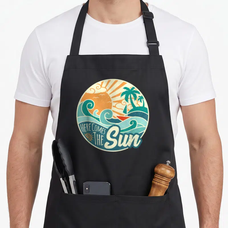 Summer come and get it apron - Retro circular print with orange sun, teal palms, waves, sailboat, and HERE COMES — shown on a black cotton canvas apron against a white background