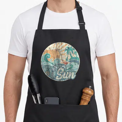 60s come and get it apron - Retro circular print with orange sun, teal palms, sailboat, and text HERE COMES THE Sun — shown on a black cotton canvas apron against a white background