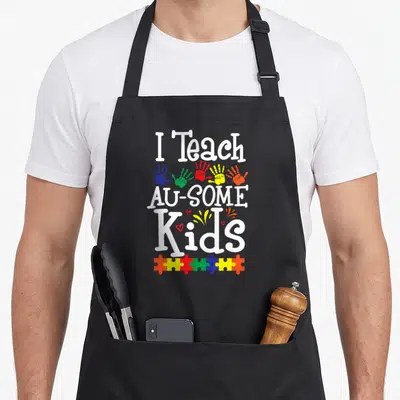Autism puzzle teacher art apron - I Teach AU-SOME Kids text with colorful handprints and puzzle piece border — shown on a black cotton canvas apron against a white background