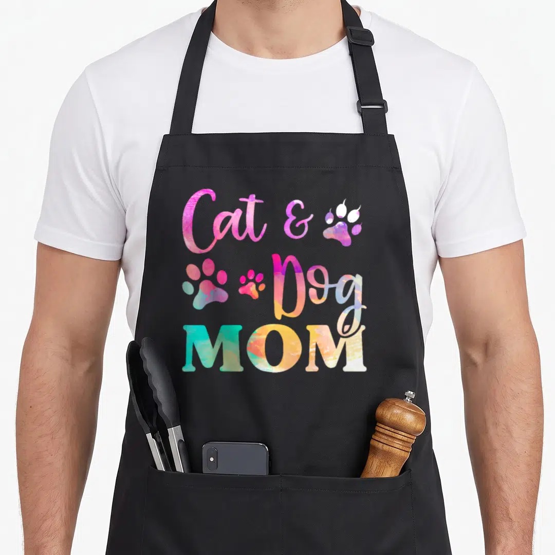 Pet dog mom apron - Stacked Cat & Dog MOM text in pink blue yellow purple with three multicolored paw prints — shown on a black cotton canvas apron against a white background