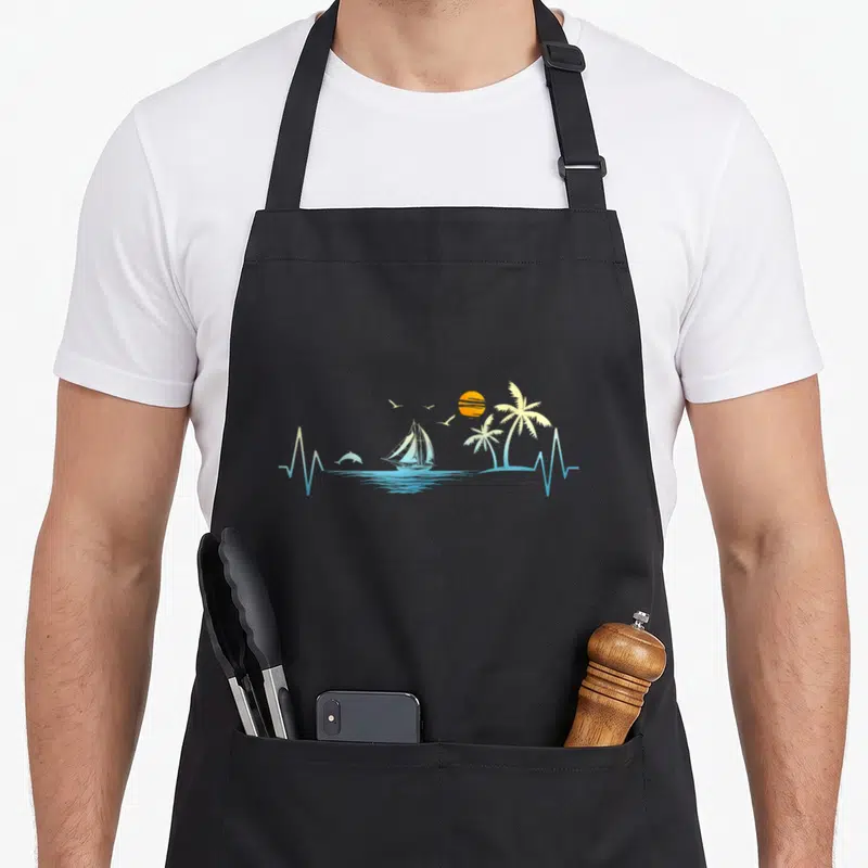 Sailboat tree apron - Sailboat dolphin palm trees sun birds in a blue and yellow heartbeat line art design — shown on a black cotton canvas apron against a white background