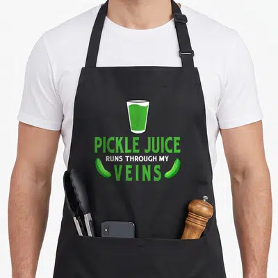 Run pickle apron - Cartoon glass of green liquid with text PICKLE JUICE RUNS THROUGH MY VEINS and two pickles — shown on a black cotton canvas apron against a white background