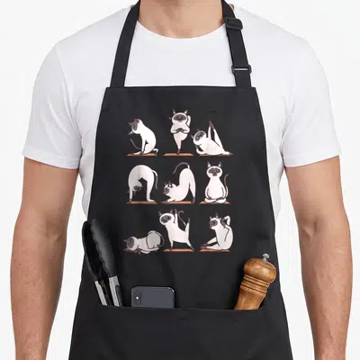 Yoga cute cat aprons - Nine Siamese cats in white grey brown doing yoga on orange mats in a grid — shown on a black cotton canvas apron against a white background