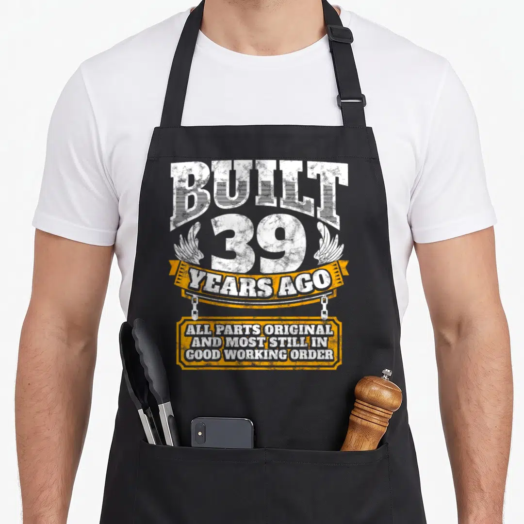 Joke birthday apron - Distressed white text BUILT 39 YEARS AGO with orange ribbon and plaque reading all parts original — shown on a black cotton canvas apron against a white background